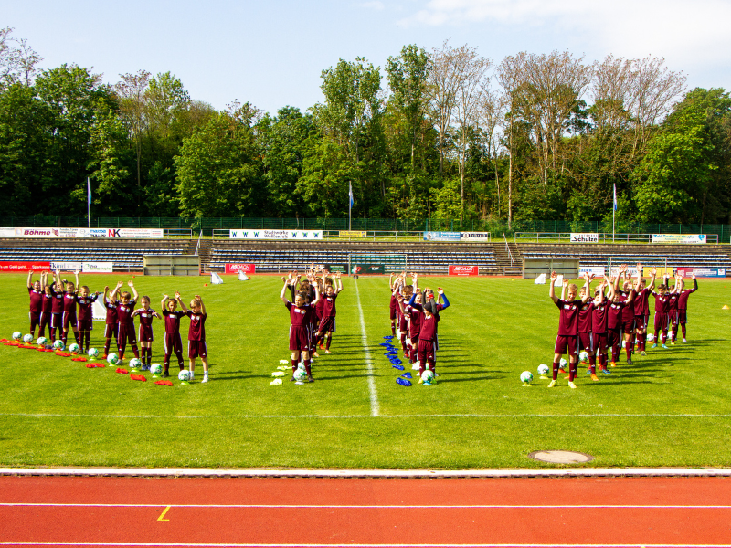 Ungefähr 30 Kinder in Fußballtrikots stehen aufgereiht auf dem Trainingsplatz und machen eine Laola